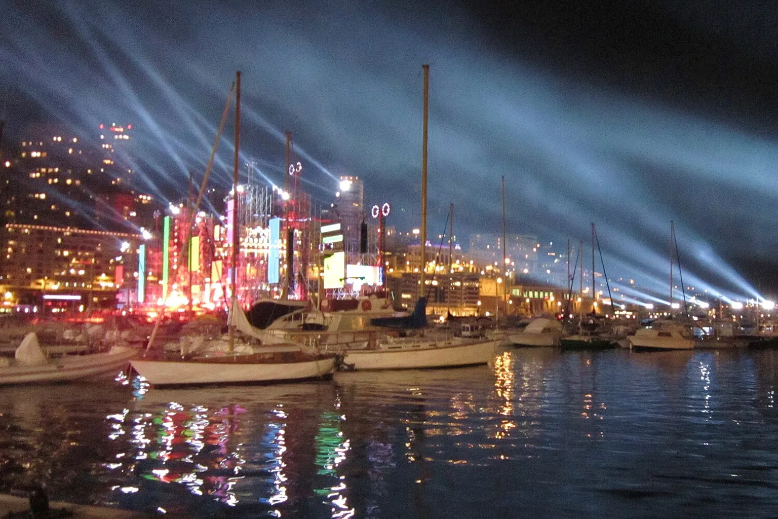 Le port de Monaco de nuit, voiliers au premier plan et faisceaux lumineux dans le ciel