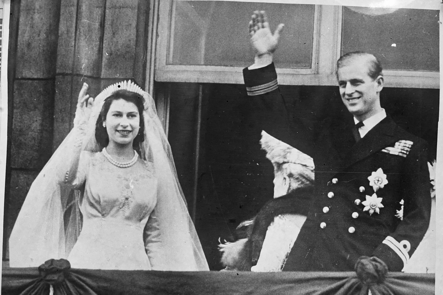 Élisabeth en robe de mariée, voile et diadème, et Philip en uniforme saluant depuis un balcon, photographie reproduite en 1947